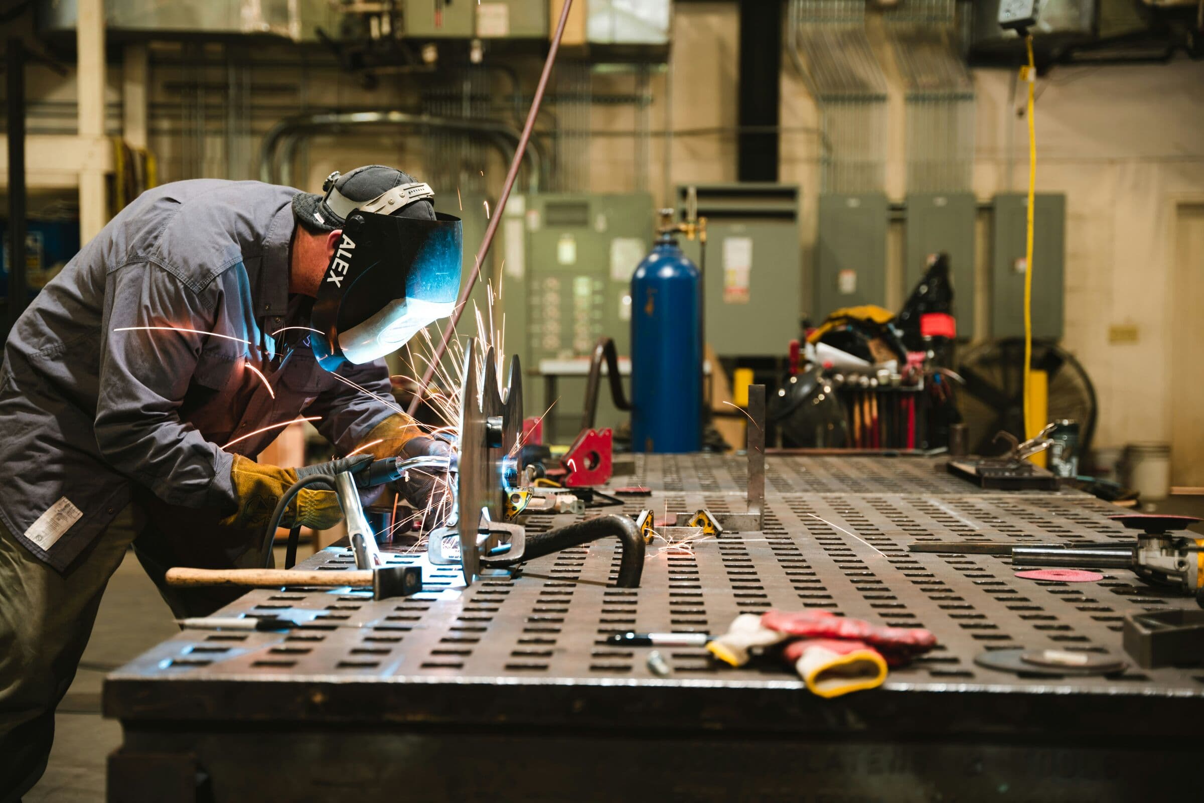 A welder in a protective mask at work on a steel fabrication table, sparks flying